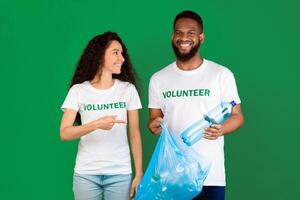 Two Diverse Volunteers Putting Wasted Plastic Bottle To Blue Garbage Bag Posing Standing On Green Background In Studio. Junk Sorting, Ecology And Volunteering Work, Keep Environment Clean Concept photo