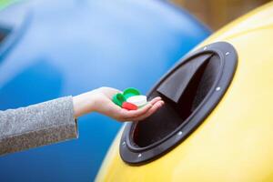 Unrecognizable Woman Throwing Handful Of Colorful Plastic Lids Bottle Caps Into Yellow Recycle Bin, Enjoying Garbage Sorting And Zero Waste Living, Cropped Image With Selective Focus, Free Space photo