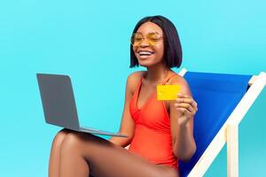 Happy African American lady using laptop in lounge chair, showing credit card, shopping online on blue studio background. Joyful black woman buying goods in web store on summer sale photo