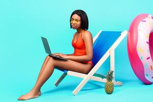 Online work during summer vacation. Unhappy black lady in swimsuit sitting in lounge chair with laptop, having to do urgent job on holidays, participating in business meeting, blue background photo