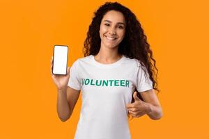Happy Volunteer Lady Showing Blank Smartphone Screen And Gesturing Thumbs-Up To Camera Posing Standing Over Yellow Studio Background. Mobile App For Charity And Volunteering Concept. Mockup photo