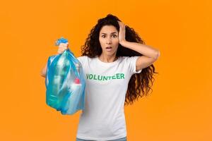 Shocked Volunteer Woman Holding Plastic Garbage Bag With Litter Looking At Camera Standing On Yellow Studio Background. Unsorted Waste And Pollution Problem, Junk Sorting Concept photo