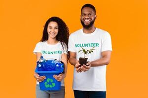 Protection Of Environment. Two Multiracial Volunteers Holding Used Plastic Bottles And Plant In A Pot Standing Over Yellow Studio Background, Smiling To Camera. Don't Litter, Care For Nature Concept photo