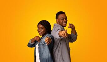 Gotcha. Joyful african man and woman pointing at camera with fingers, cheerful black millennial couple indicating somebody, having fun while standing isolated over yellow background, free space photo