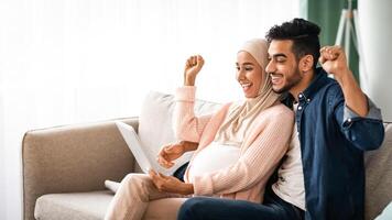 Excited Pregnant Muslim Couple Celebrating Success With Digital Tablet At Home, Happy Expectant Middle Eastern Spouses Looking At Tab Computer Screen, Shaking Fists And Exclaiming With Excitement photo