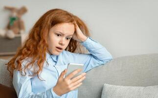 Shocked Surprised Young Teen Girl With Cellphone Reading Bad News Message Touching Head In Amazement Sitting On The Couch At Home. Confused Mobile Phone User, Call From Unknown Number photo
