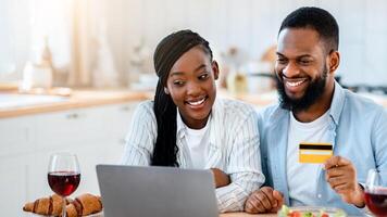 Food Ordering. Happy Black Spouses Using Laptop And Card In Kitchen, Making Online Delivery Order With Computer, Sitting At Table At Home And Enjoying Internet Purchases, Free Space photo