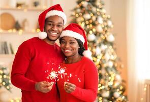 Happy New Year and Merry Xmas at home together. Smiling young african american guy and lady in Santa Claus hats hold sparklers, hug and look at camera in living room interior with tree and garlands photo