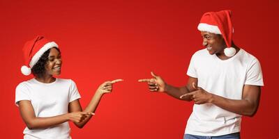 Emotional african american young man and woman in Santa hats pointing at copy space on orange studio background, showing advertisement for New Year or Christmas deal, offer, sale, discount photo