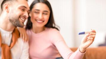 Positive Pregnancy Test. Joyful Arab Couple Hugging Awaiting Baby And Celebrating Result Sitting On Sofa At Home. Childbirth, Family Happiness. Happy Parents-To-Be Concept. Selective Focus photo