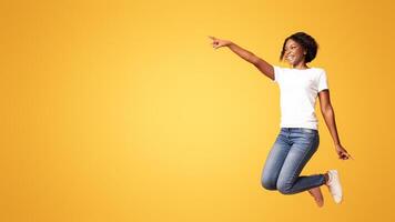 Excited black woman jumping up and showing copy space over orange studio background. Emotional african american millennial lady smiling and pointing at advertising or announcement photo