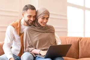 Modern Muslim Spouses Using Laptop Watching Movie Together Sitting Embracing On Couch At Home On Weekend. Lovely Arab Couple With Computer Relaxing Indoors. Family Gadgets And Technology photo