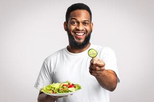 Healthy Eating. Cheerful African Guy Posing With Vegetable Salad Offering Slice Of Cucumber To Camera Standing Over White Studio Background. Try Vegan Diet, Male Nutrition And Weight Loss Concept photo