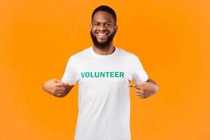Happy african american volunteer guy showing his uniform t-shirt with text, smiling to camera posing on yellow background, studio shot. Portrait of joyful activist. I choose volunteering career photo
