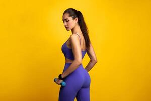 Fitness Instructor. Portrait Of Confident Young Woman Exercising With Two Dumbbells Posing Isolated Over Orange Background, Standing Back To Camera. Studio Shot, Copy Space, Banner For Fitness Club photo