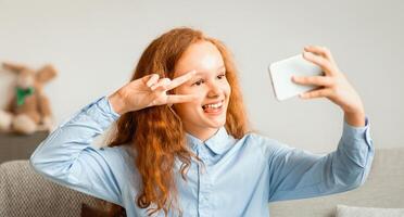 Portrait of cool cheerful red-haired teenage girl taking selfie for social network, holding smartphone in hand shooting picture on front camera, showing v sign gesture, sitting on couch at home photo