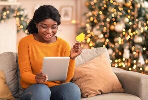 Modern purchases during self-isolation at home. Smiling african american millennial female uses tablet and credit card to shop online in living room interior with Christmas tree and shining garlands photo