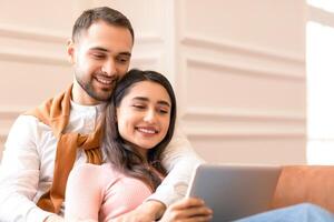 Happy Modern Arab Couple Using Digital Tablet Computer Watching Movie Online Sitting On Sofa At Home. Spouses Browsing Internet Together Indoors. Family Weekend Concept photo