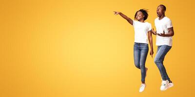 Excited african-american couple jumping up and cheerfully pointing away at empty space, orange studio background. Emotional young black man and woman aiming at advertisement, full length photo