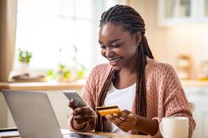 Online Payments. Cheerful black lady using smartphone and credit card in kitchen at home, smiling african woman sitting at table with laptop, paying bills in internet or transferring money via app photo