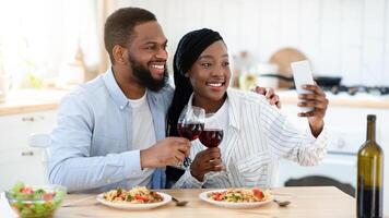 Happy Moments. Cheerful Black Couple Taking Selfie On Smartphone While Having Romantic Date At Home, Young African American Spouses Sitting At Table In Kitchen And Clinking Glasses With Red Wine photo