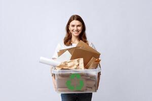 Recycling And Waste Sorting Concept. Smiling Young Woman Holding Plastic Box With Paper Garbage, Standing Over Light Background In Studio, Portrait Of Millennial Eco-Friendly Lady, Copy Space photo