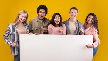 Group of friends laughing, sharing good and positive mood. Happy young multiracial students with open mouths hold banner with blank space, isolated on orange background, studio shot, panorama photo