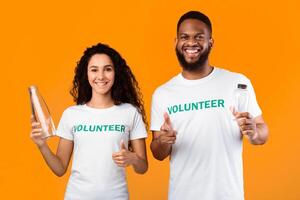 No More Plastic. Two Volunteers Holding Glass And Plastic Bottles Pointing Fingers At Camera Standing In Studio On Yellow Background. Make The Right Choice, Stop Using Single-Used Bottle photo