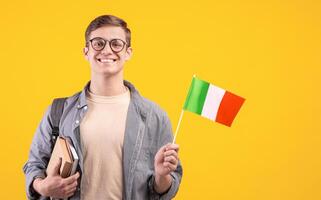 Love of country, education and travel, exchange student and new pupil. Young smiling guy in glasses with backpack holds books and small flag of Italy, isolated on beige background, studio shot photo