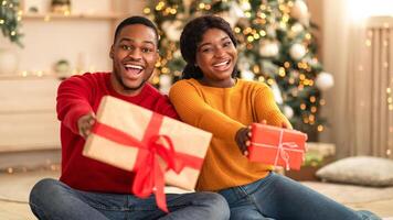 Fulfillment of wishes in New Year at home during lockdown. Laughing millennial african american man and woman show boxes with gifts and ribbons in living room interior with Xmas tree and garlands photo