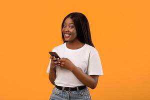 Excited black lady holding brand new smartphone and smiling, recommending new awesome mobile application, orange studio background, copy space. Smartphone, mobile phone, cellphone concept photo