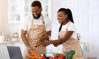 Happy African American Spouses Using Laptop Computer While Cooking In Kitchen Together, Searching Recipe In Internet, Smiling Black Couple Preparing Healthy Food For Dinner At Home, Copy Space photo