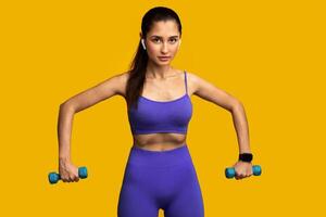 Sport And Fitness Concept. Portrait Of Confident Young Strong Woman In Wireless Headphones Exercising With Two Blue Dumbbells, Listening To Music, Posing Isolated On Orange Studio Background photo