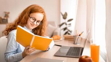 Learning And Education Concept. Portrait of young smiling red-haired girl wearing eyeglasses, sitting at desk with laptop, reading and doing homework, holding notebook. Teen studying at home photo