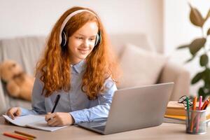 Doing Homework. Smiling young teen girl sitting at desk with pen, writing and taking notes in notebook, using and looking at laptop computer. Cheerful pupil in headphones watching online course photo