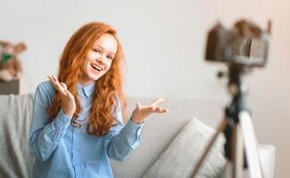 Blogging Concept. Smiling red-haired teen girl recording lifestyle blog talking to camera on tripod, sitting on the couch at home. Happy young influencer filming review for her internet channel photo