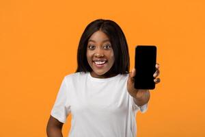 Mockup for application screen or text. Smartphone with empty screen in cheerful black lady hand, orange studio background, closeup. African american pretty woman showing blank mobile phone screen photo