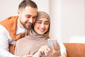 Happy Muslim Couple Using Smartphone Texting And Browsing Internet Sitting On Couch At Home. Family Leisure. Cheerful Arab Spouses Watching Online On Phone. Great Mobile Application Concept photo