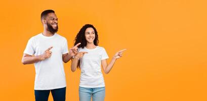 Multiracial Couple Pointing Fingers Aside Showing Copy Space For Text Standing Over Yellow Studio Background. Look There Concept, Advertisement Banner. Panorama photo