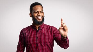 Look At This. Cheerful Black Millennial Guy Holding Invisible Object On Finger Or Pointing Upward Standing Over White Studio Background. African Guy Posing Advertising Your Product photo