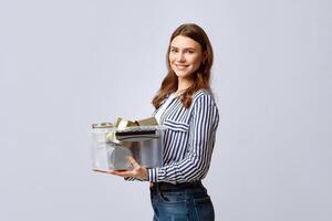 A young woman stands indoors, smiling broadly while holding a transparent container filled with recyclable materials. She showcases a commitment to eco-friendly practices and sustainability. photo