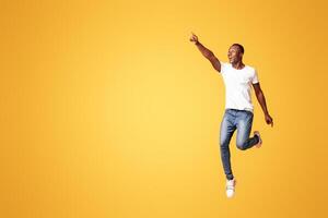 Emotional black man jumping and pointing at copy space for advertising or announcement on orange background, studio full length shot. Cheerful african american guy showing great advertisement photo