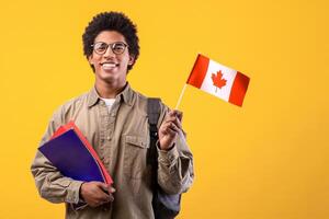Education, school, college and university. Happy young african american guy in glasses holding notepads and small Canada flag and ready to study, isolated on orange background, free space, studio shot photo