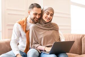Modern Muslim Couple Watching Movie Online On Laptop Computer Sitting And Hugging On Couch, Relaxing At Home On Weekend. Lovely Arab Spouses Browsing Internet Together photo