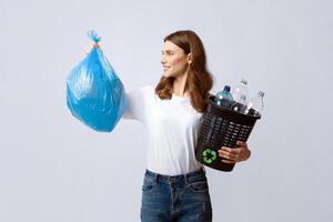 Waste Sorting. Positive Young Lady Holding Bag With Garbage And Bucket Filled With Plastic Bottles, Millennial Woman Enjoying Eco-Friendly Living, Standing Over Grey Studio Background, Copy Space photo