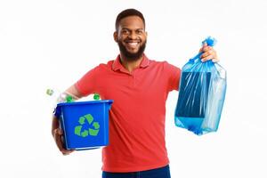 Black Man Holding Box With Recycling Plastic Litter And Blue Garbage Bag Standing On White Studio Background, Smiling To Camera. Waste Sorting And Recycling Concept photo