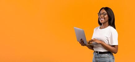 Happy african american young woman with glasses holding newest laptop and smiling, orange studio background, panorama with copy space. Cheerful black lady studnet using notebook, studying online photo