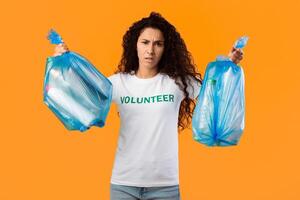 Displeased Female Volunteer Holding Two Plastic Garbage Bags Full Of Litter And Wasted Bottles Posing On Yellow Studio Background. Junk Sorting, Ecology Concept photo