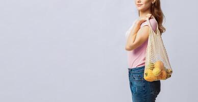 A woman is seen holding a reusable bag filled with fruits, showcasing her commitment to eco-friendly practices. She stands confidently against a simple background, copy space photo