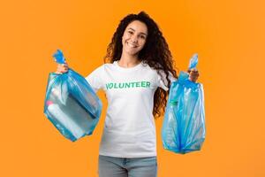 Sort Your Waste. Volunteer Lady Holding Blue Plastic Garbage Bags Posing Smiling To Camera Standing In Studio Over Yellow Background. Litter And Trash Sorting, Clean Environment Concept photo